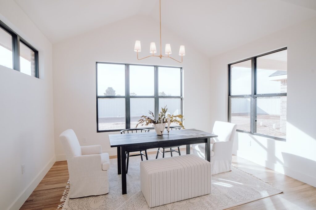 Minimalist dining room with large windows, a black table, white upholstered chairs, a striped ottoman, light fixture, and decorative plants on the table. airbnb cleaning